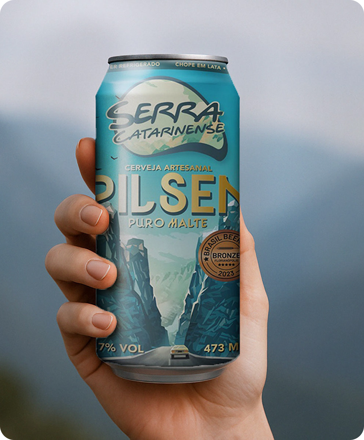 Mano sosteniendo una lata de la Pilsen Serra Catarinense, ilustrada con un paisaje de carretera de montaña.